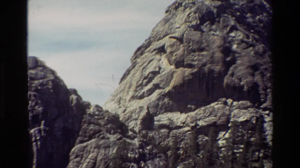 1981 8mm frame of a rugged rocky peak with smaller cliffs, pale sky, and visible film gate borders.