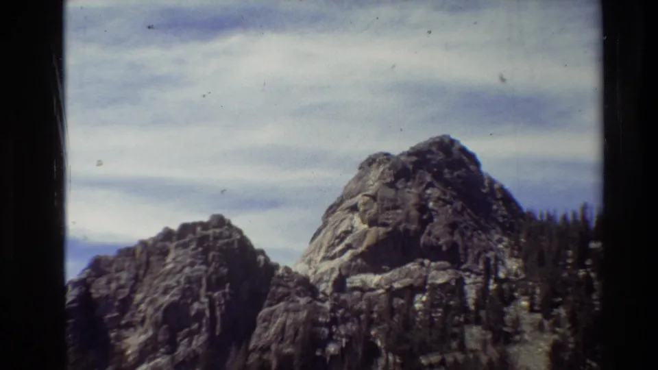 1981 8mm frame showing two rugged rocky peaks, evergreen forest at right, and a pale clouded sky.