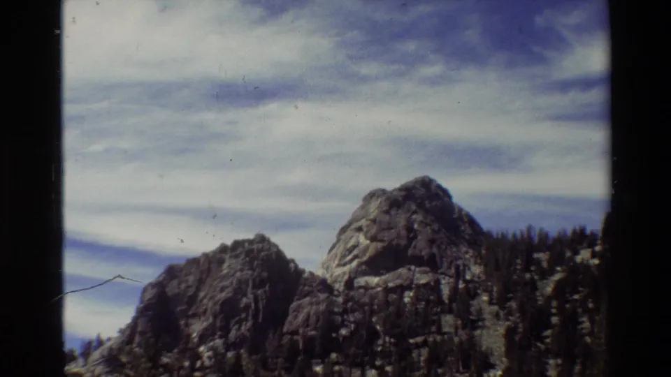 Wider 1981 home-movie view of rocky mountain peaks with evergreen trees on the slope and visible 8mm wear.