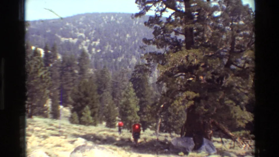 A handheld 8mm view shows two hikers with red packs walking through pine forest toward distant mountain ridges.