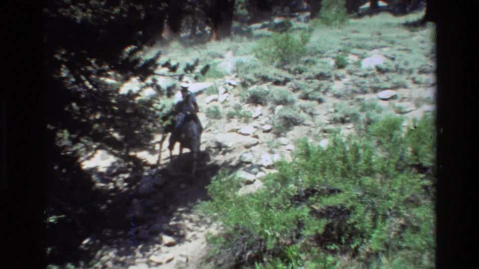 A distant horseback rider moves along a rocky, partly shaded trail in a wooded outdoor area, from 1981 home-movie footage.