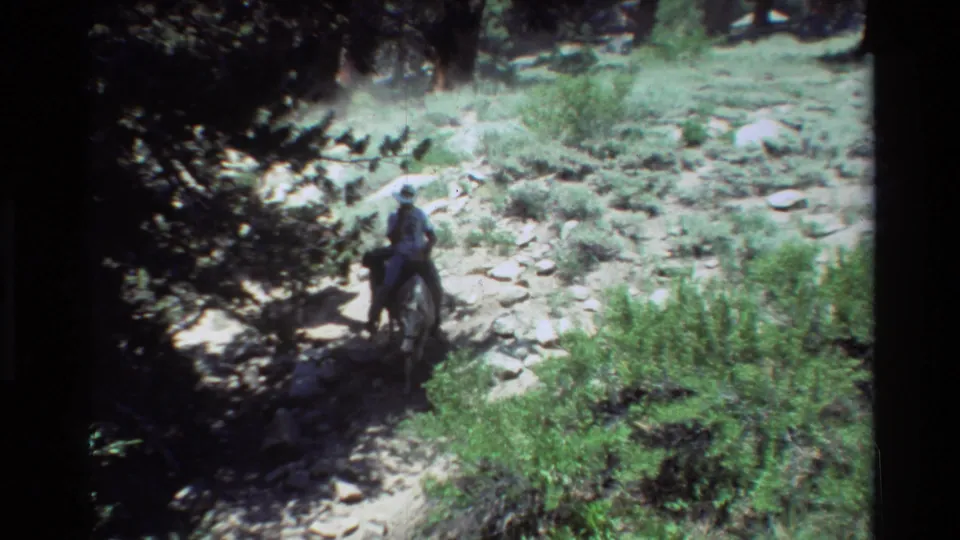 1981 8mm frame of a brimmed-hat rider on horseback leading pack animals along a rocky, shrub-lined trail.