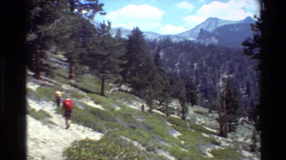 Distant hikers with colorful packs move through a forested alpine slope, with mountains visible in the background.