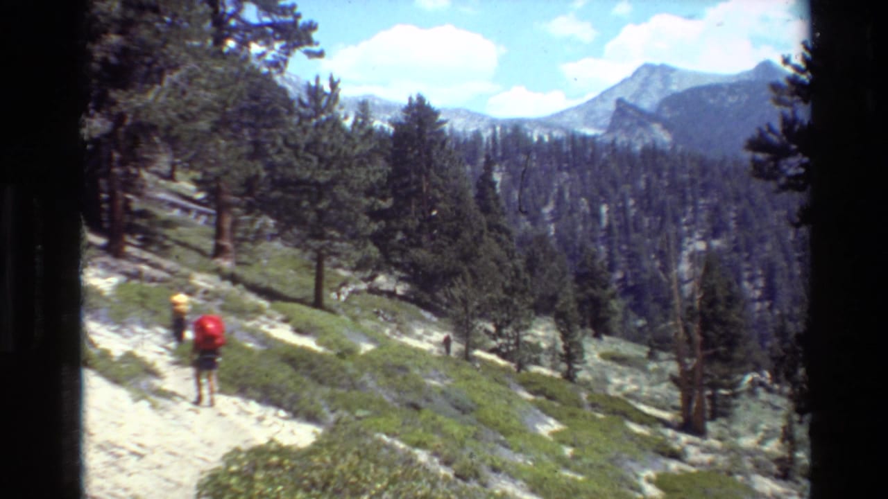 1981 8mm home-movie frame of a distant backpacked hiker on a mountain trail among evergreens, with peaks beyond.
