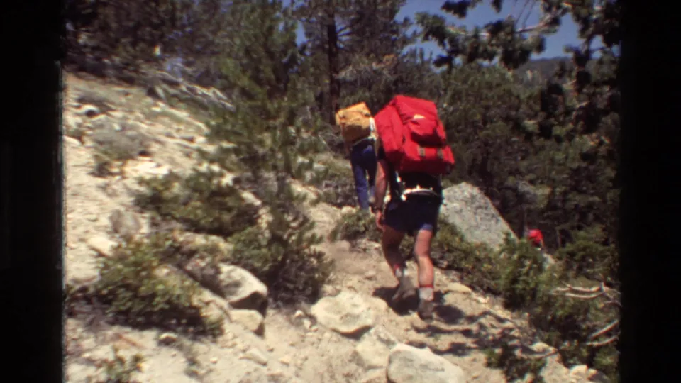 Two hikers seen from behind on a rocky forest trail, with one carrying a bright red backpack in the 1981 8mm clip.