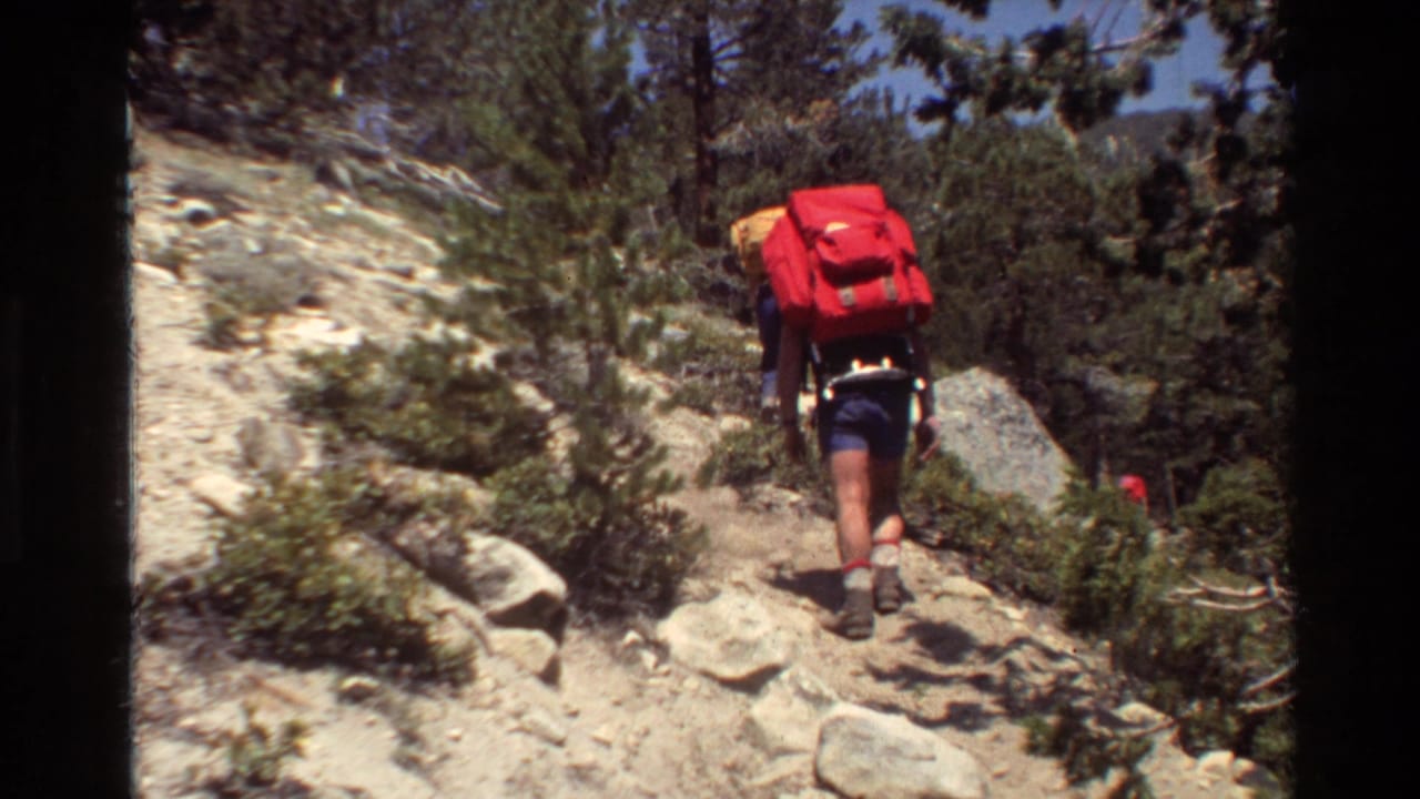 A hiker in shorts carries a red backpack on a rocky forest trail in 1981 home-movie footage.