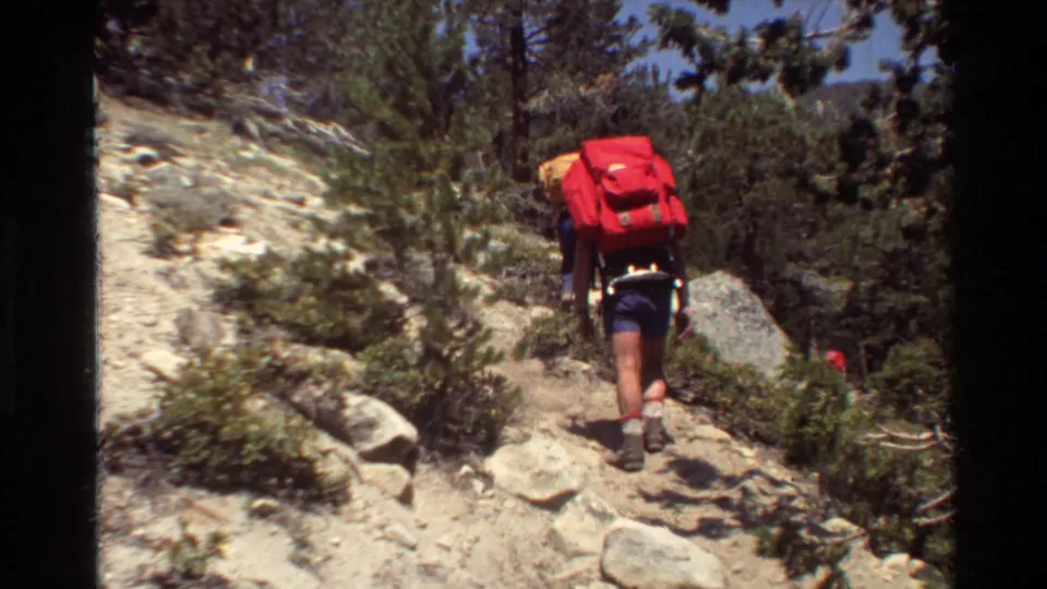 A hiker in shorts carries a red backpack on a rocky forest trail in 1981 home-movie footage.