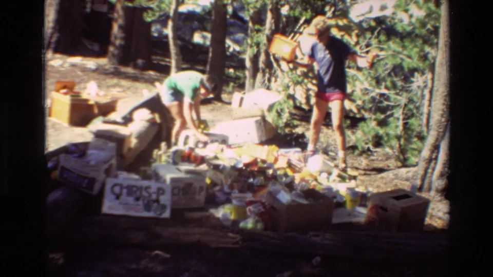 Two people work among cardboard boxes and scattered containers in a forested mountain area.