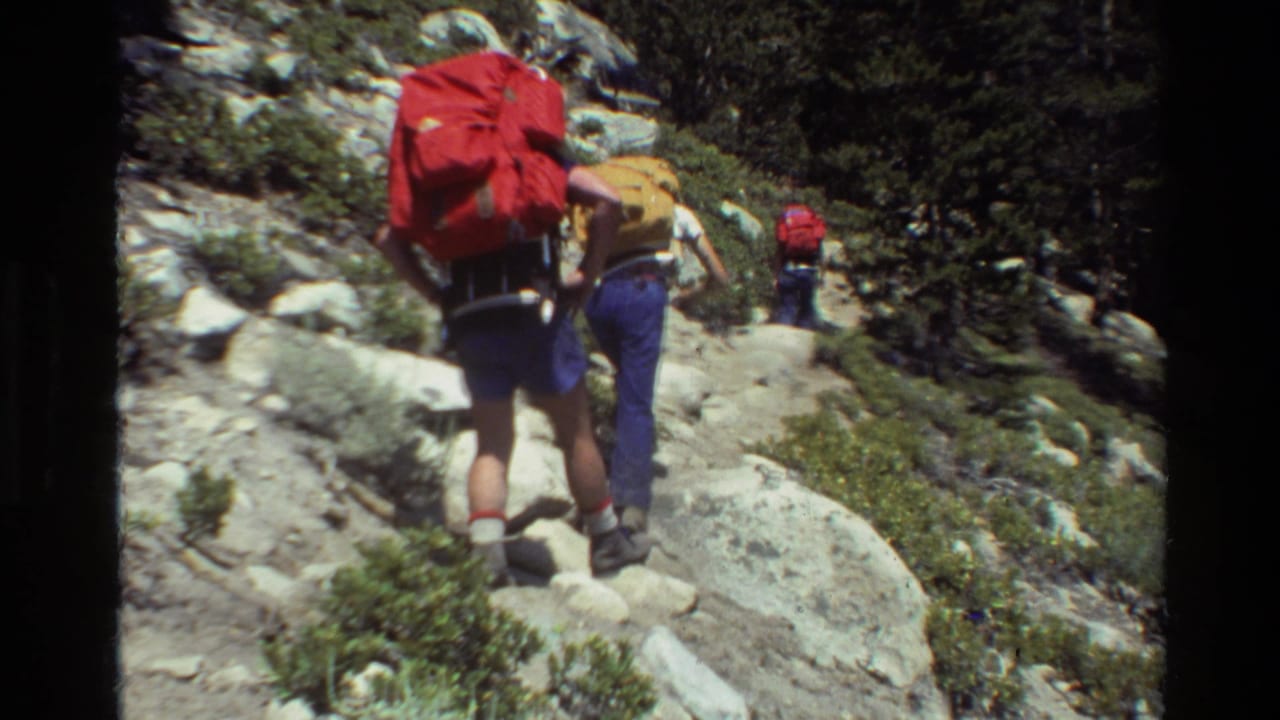 Hikers with red and yellow backpacks walk uphill on a rocky trail.