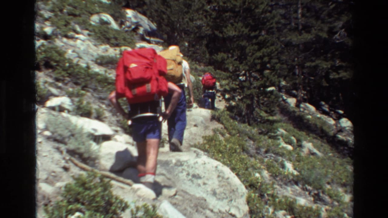 Backpackers continue along a rocky trail bordered by trees and boulders.