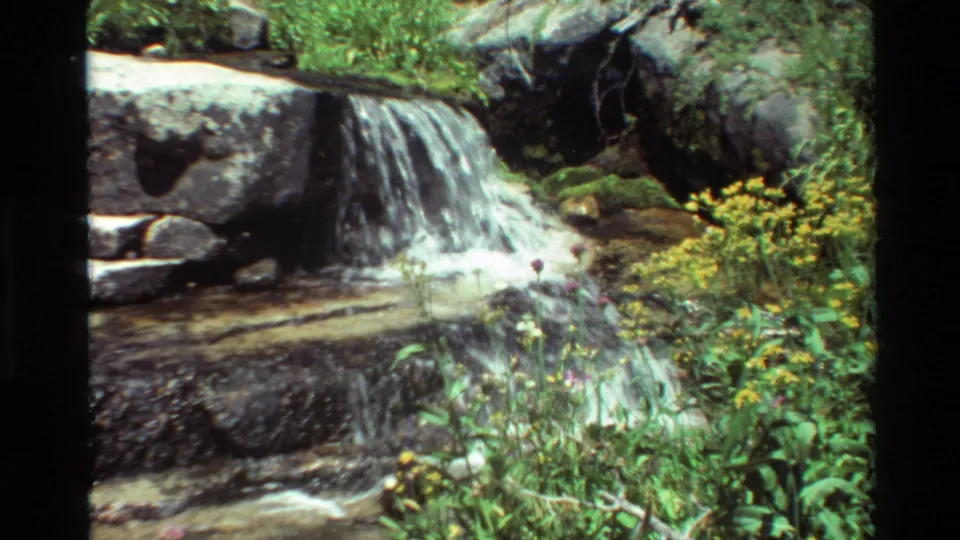 8mm home-movie frame of a small creek waterfall, rocks, and green vegetation after the hiking trail views.