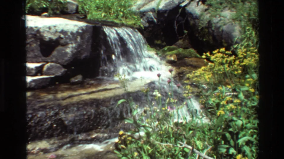A close 8mm view of a small waterfall over rocks, with stream water and vegetation in a mountain setting.