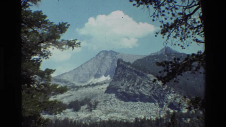 1981 8mm home-movie frame showing a mountain range in British Columbia, framed by pine branches under a bright cloud.