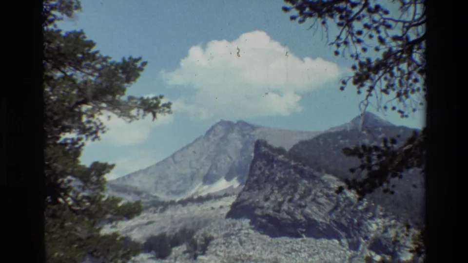 1981 8mm home-movie frame showing a wide mountain landscape with tree branches, rocky terrain, and a bright cloud overhead.