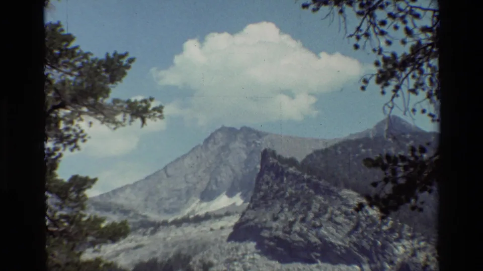 1981 8mm home-movie frame showing the mountain landscape from a tree-framed viewpoint, with rocky slopes and a large cloud.