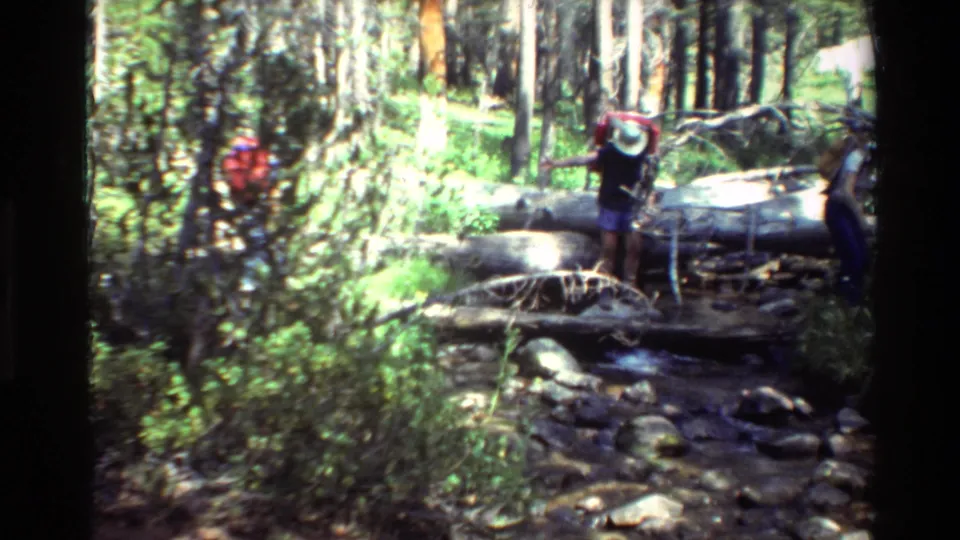 A hiker carrying a red backpack steps across a fallen-log crossing as another person waits near the creek in vintage 8mm forest footage.