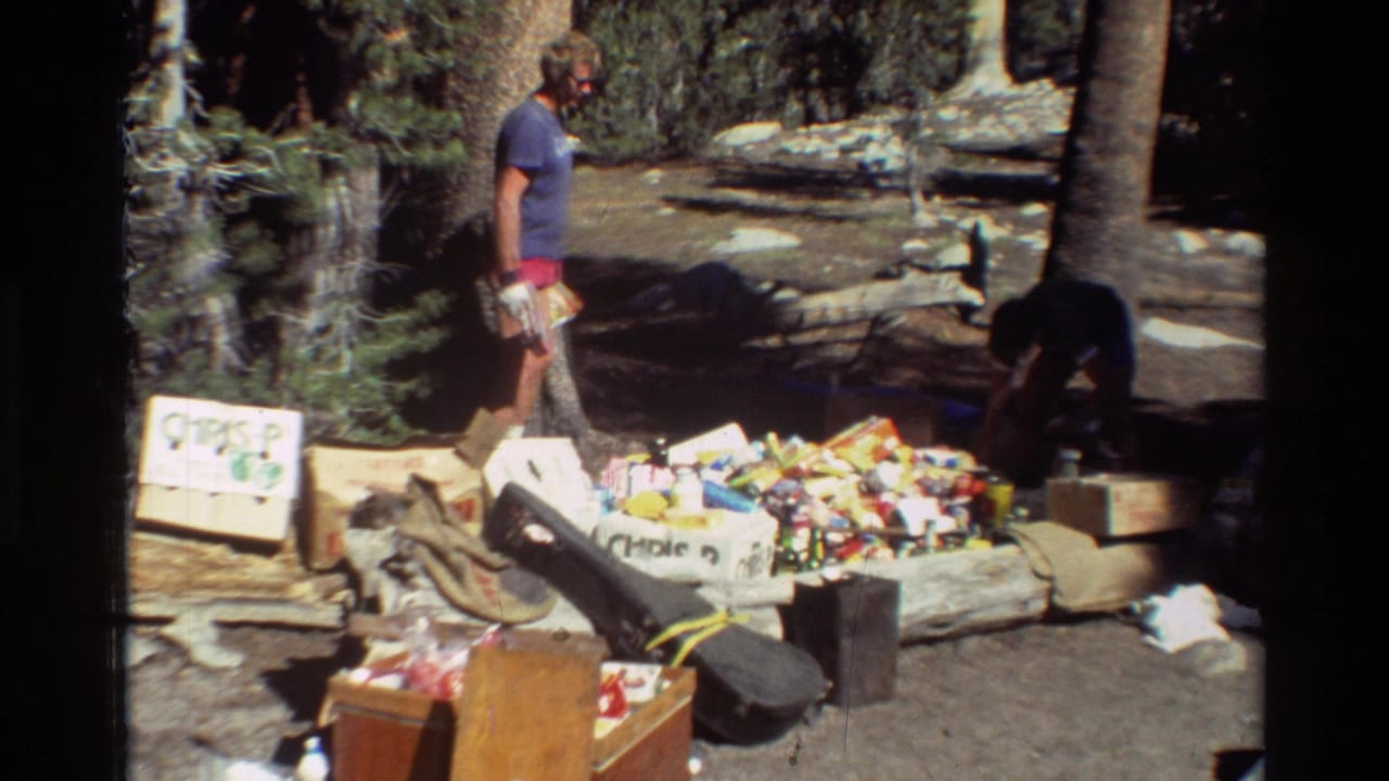 Man stands near camp supplies and boxes in a wooded campsite.