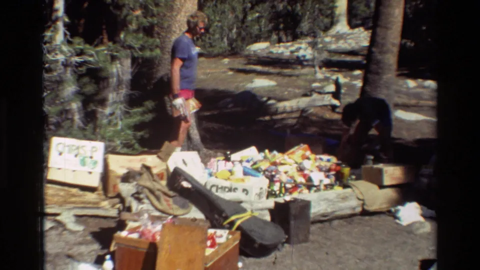 In metadata-listed 1981 British Columbia footage, a man in a blue shirt and red shorts stands beside camp supplies, boxes, and a guitar case in forest light.