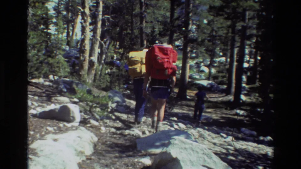 Two hikers with large backpacks walk away along a rocky trail through tall trees in the metadata-listed British Columbia home movie.