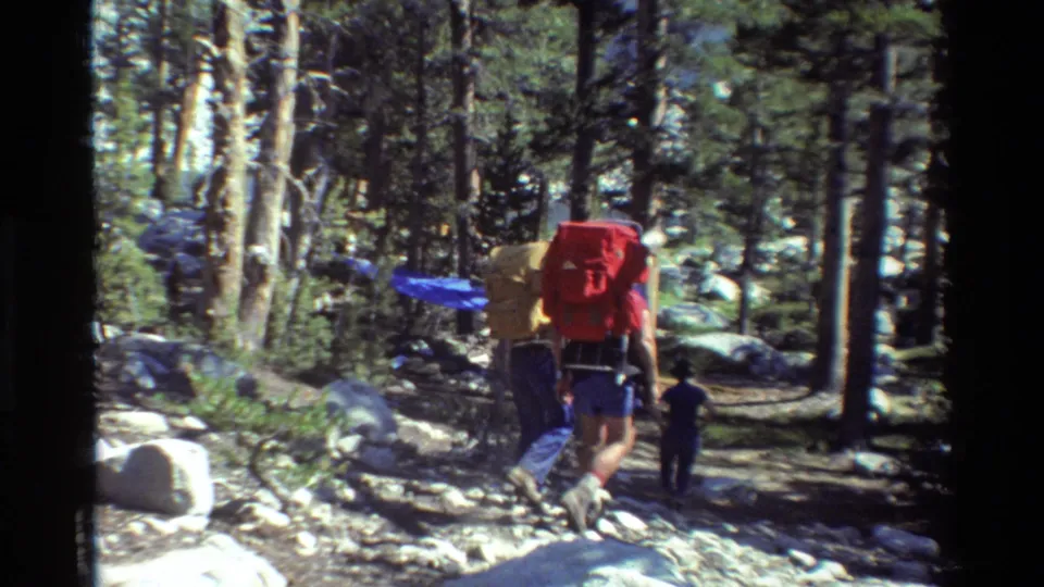 The hikers continue away from the camera through a rocky wooded trail, carrying bright packs in handheld 8mm footage.
