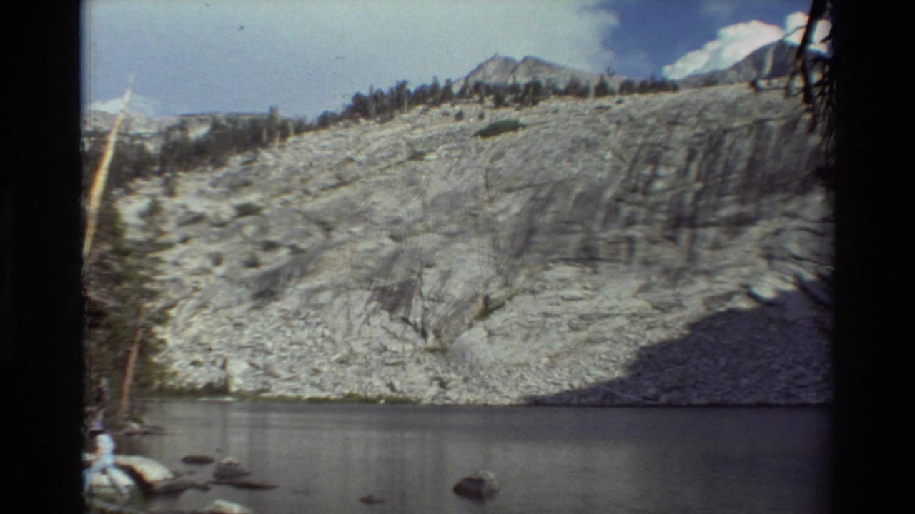 1981 8mm frame of a pale rocky mountainside rising above still water, with distant peaks, trees, and visible film edge shadows.