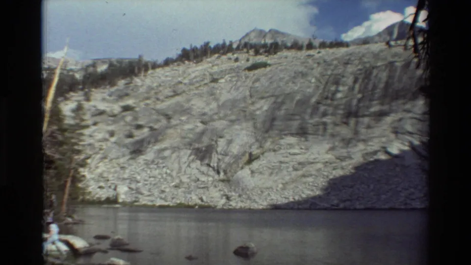 1981 8mm frame of a pale rocky mountainside rising above still water, with distant peaks, trees, and visible film edge shadows.