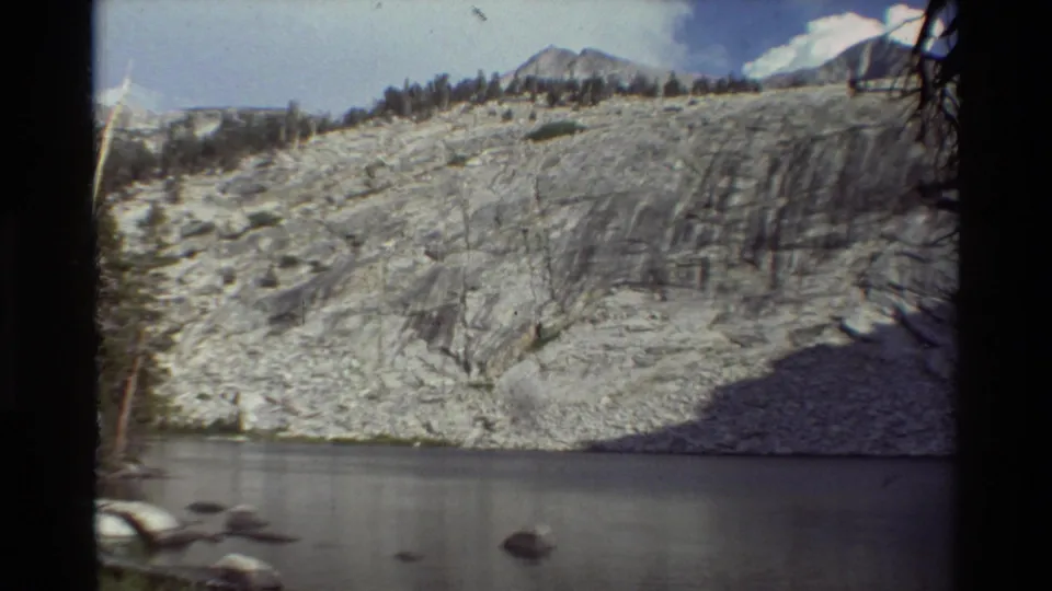 Metadata-dated 1981 8mm view across quiet water toward a broad rock face, scattered trees, and distant mountain peaks.