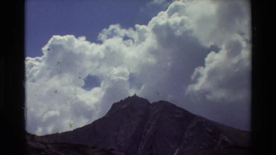 1981 8mm upward view of a jagged mountain peak beneath bright cumulus clouds, with visible grain and gate-edge shadow.