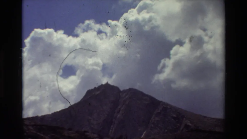 Metadata-dated 1981 8mm frame of a rugged summit under towering clouds, with scratches, dust specks, and soft color fading visible.