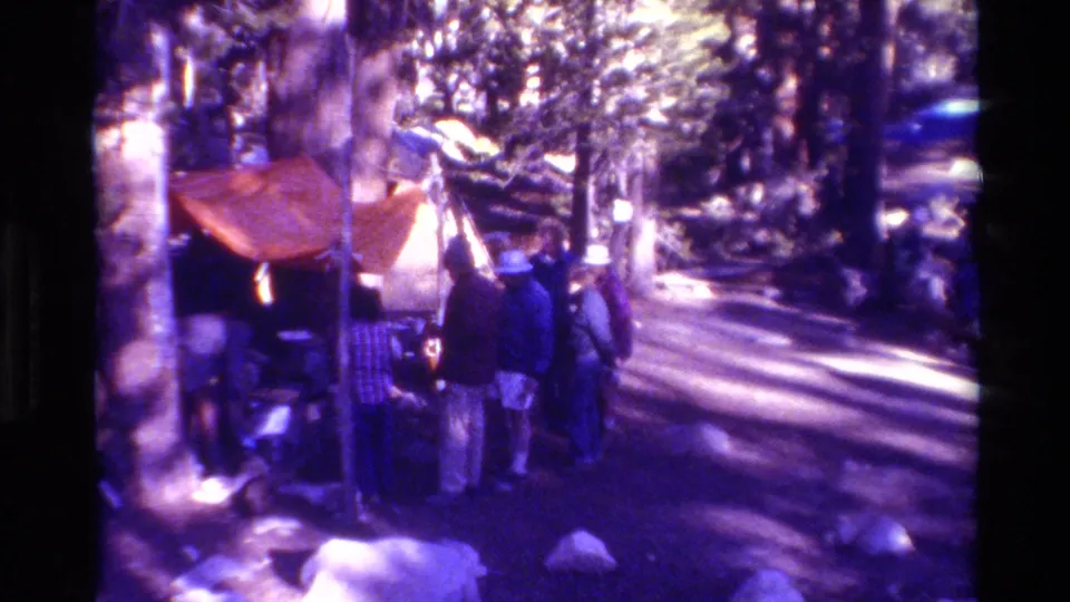 Adults in casual outdoor clothing stand by an orange tent at a wooded campsite, with rocks and tall trees around them.