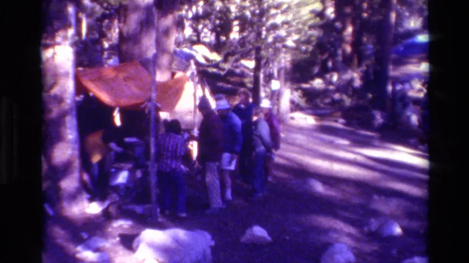 A wooded campsite scene shows people beside an orange tent, with tall trees and dappled sunlight across the ground.
