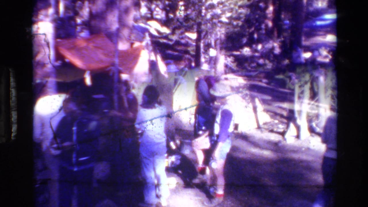 Adults at a wooded campsite beneath an orange tarp, with trees and camping gear nearby.
