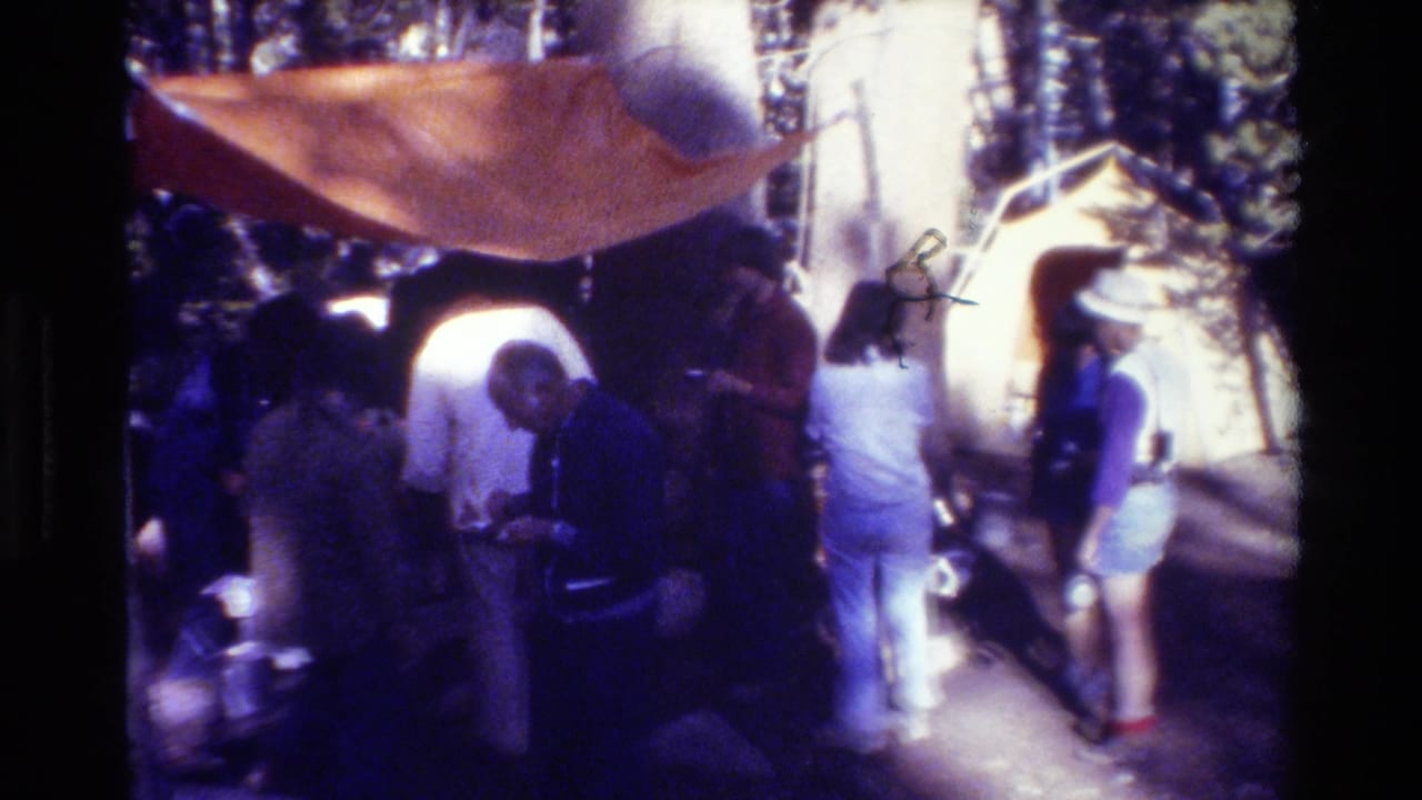 Group clustered under a large orange tarp at a wooded campsite.