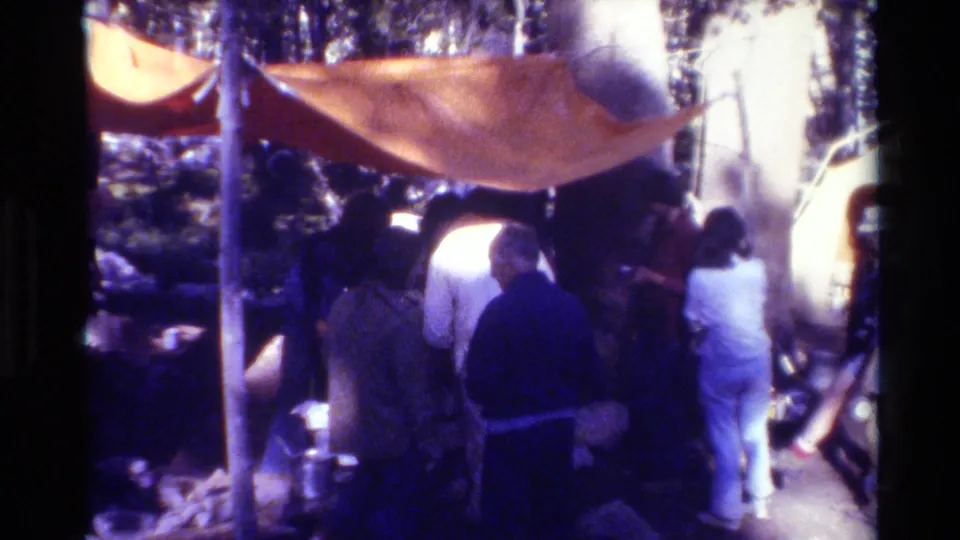 8mm home-movie frame of adults at a British Columbia forest campsite, gathered under an orange tarp among trees and camp equipment.