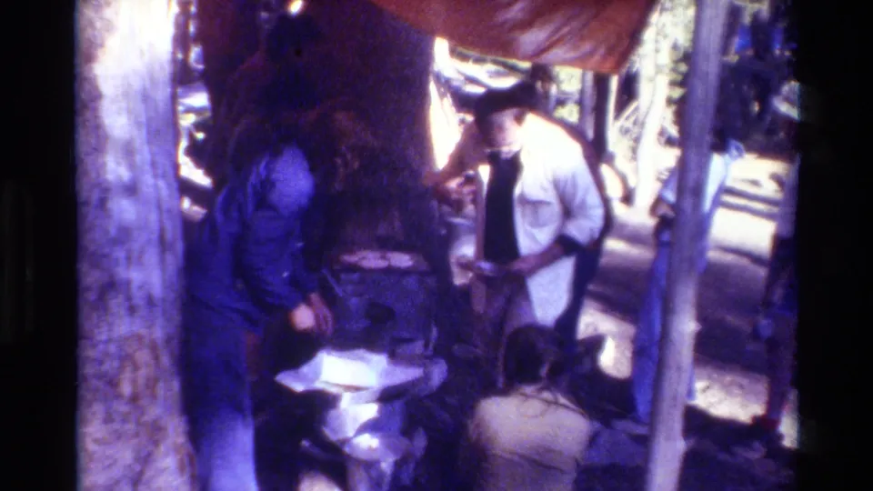 Adults at a wooded campsite in 1981 footage, with a tree trunk, tarp edge, seated figure, and camping items in soft 8mm color.