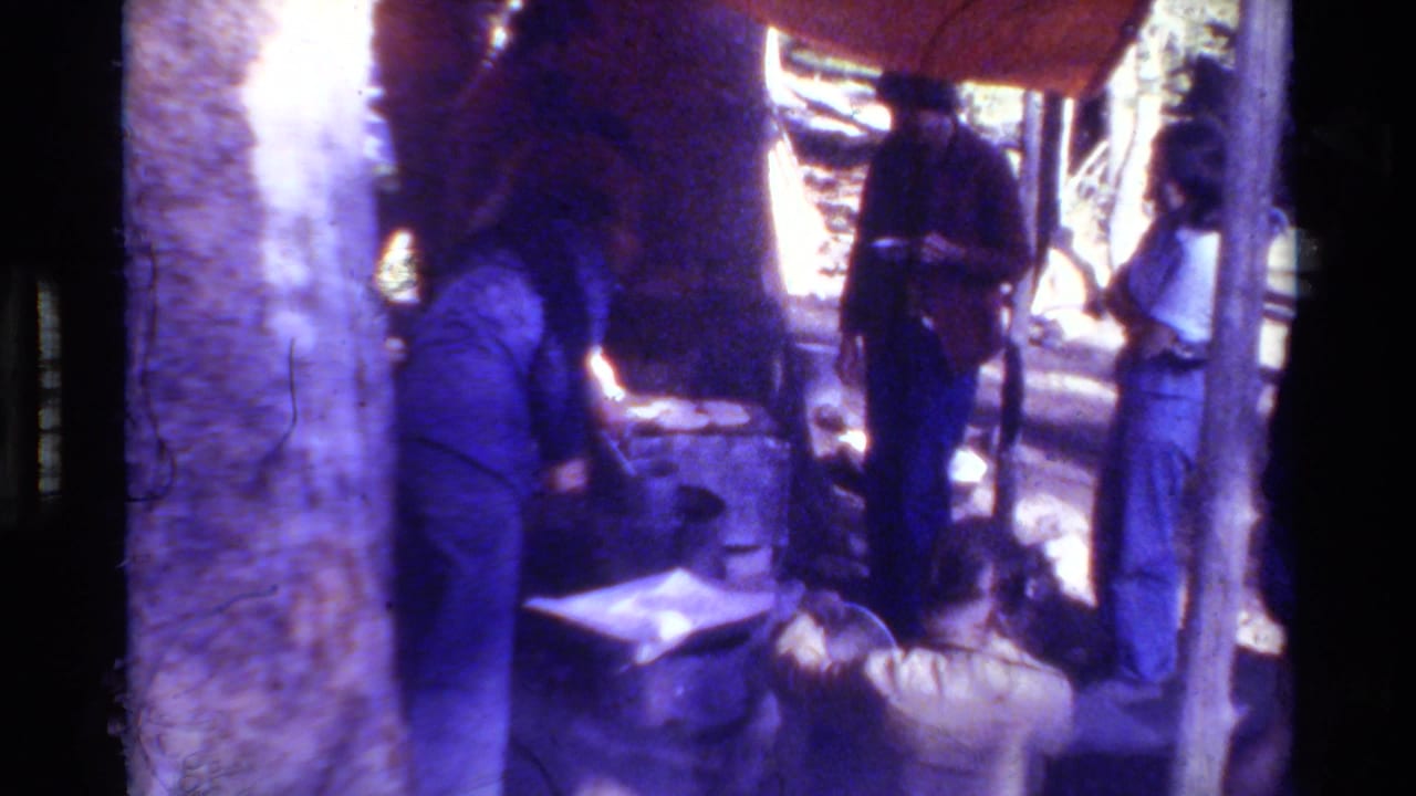 1981 8mm home-movie frame showing several casually dressed people around outdoor cooking gear in a wooded British Columbia-listed setting.