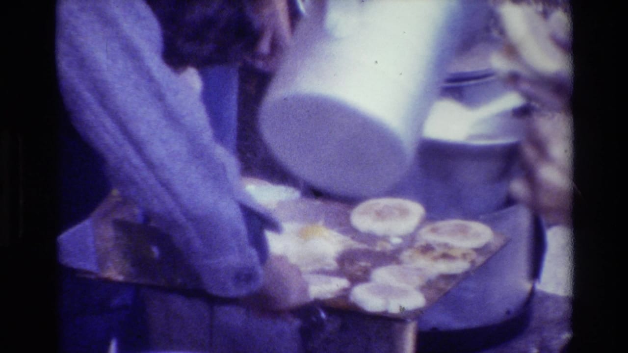 Hands work over a griddle with round food pieces while a white container is held nearby.