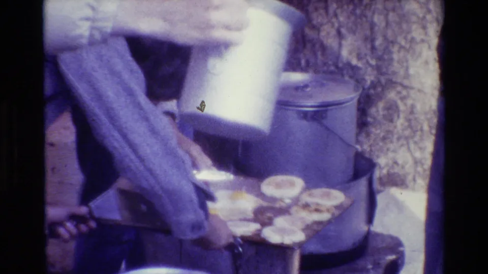 A person pours or tips from a white container near a griddle, with a large pot and round food pieces visible in the outdoor cooking setup.