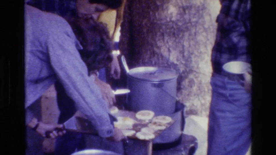 Casually dressed people cluster around a large pot and griddle with round food pieces in a wooded 1981 home-movie cooking scene.