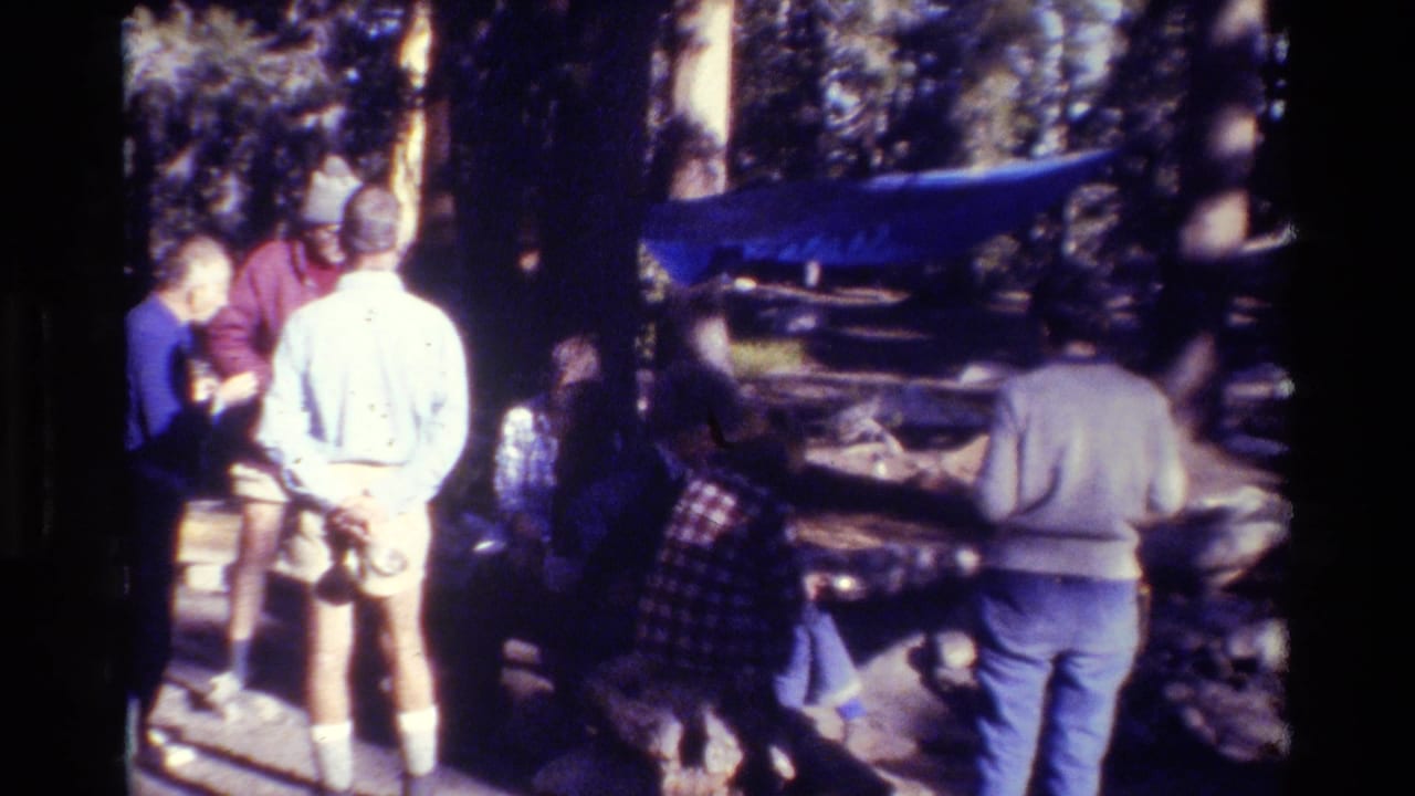 1981 8mm frame of bundled campers gathered in a wooded campsite with a blue tarp visible between trees.