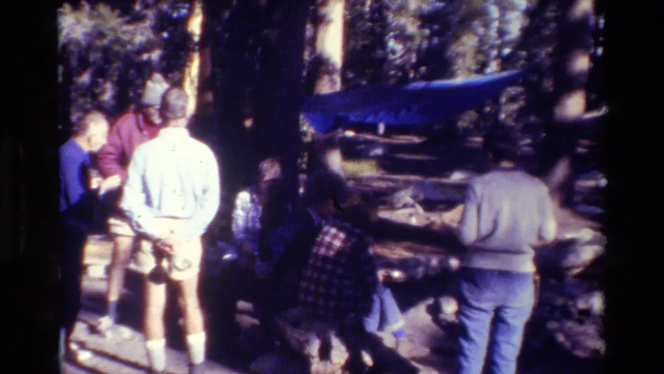 1981 8mm frame of bundled campers gathered in a wooded campsite with a blue tarp visible between trees.
