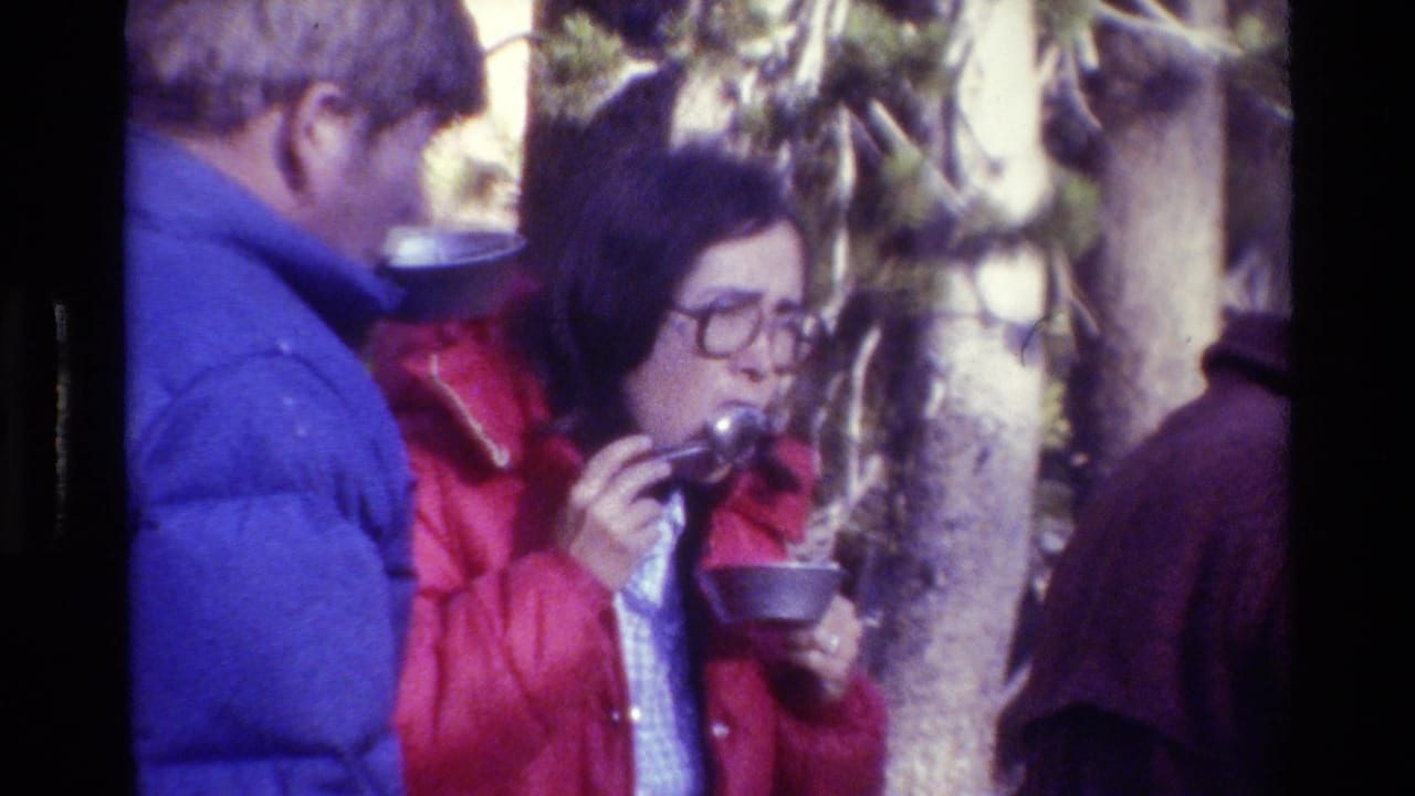 Person in a red jacket eating from a metal bowl beside another camper.