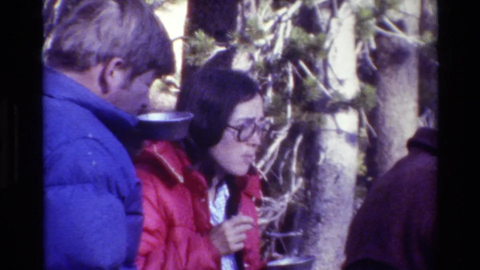 1981 8mm frame showing two bundled campers with metal bowls and spoons during a forest campsite meal.