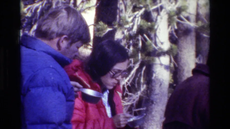 1981 wooded campsite frame with a camper in a red jacket holding a metal bowl and spoon beside another bundled person.