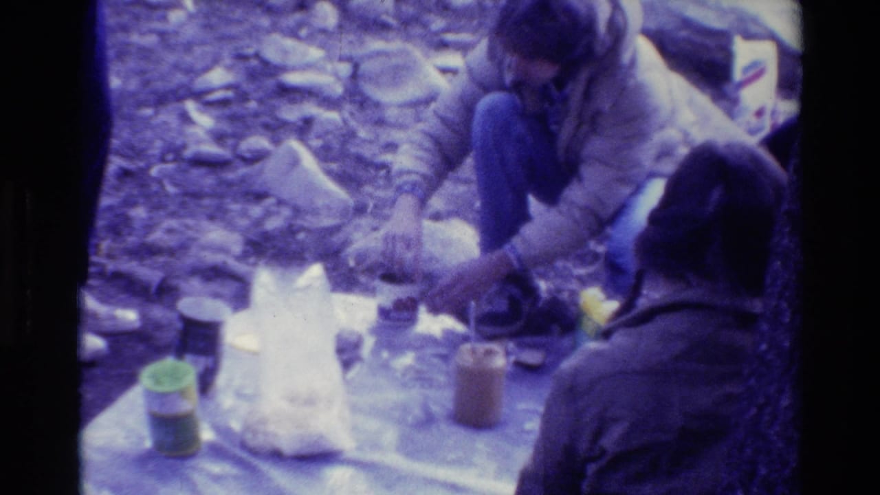 Campers crouch around food and containers on a blanket in a rocky wooded area.