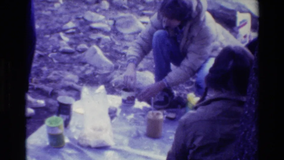 1981 8mm home-movie frame of campers preparing food on a blanket in a rocky wooded campsite.