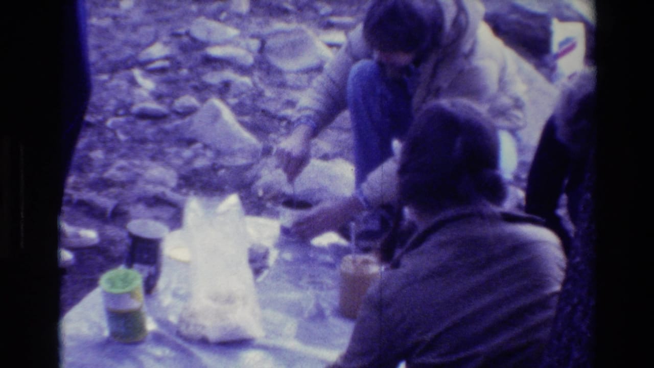 Two campers sit near a picnic blanket with jars and a plastic bag on rocky ground.