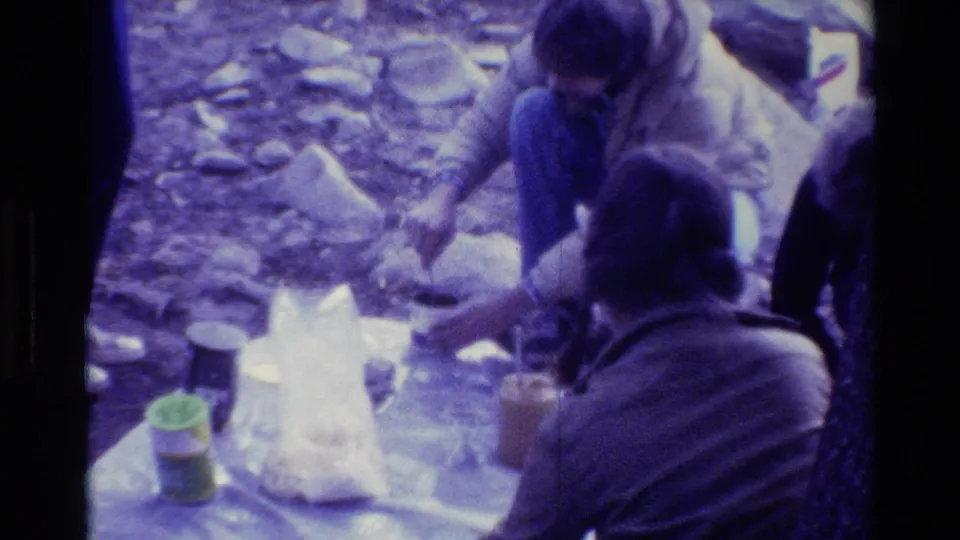 Campers sit beside food containers and a white plastic bag in a rocky wooded campsite, filmed on 8mm in 1981.
