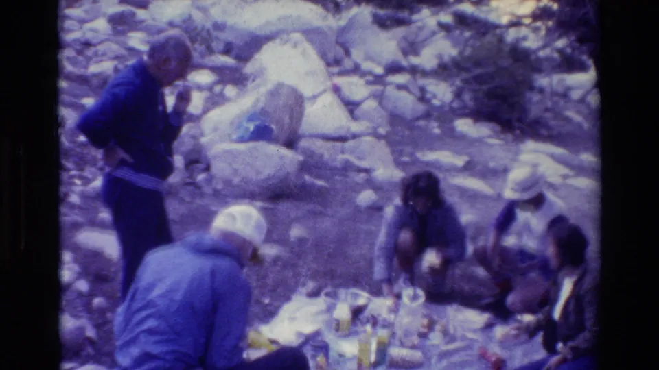 Group of campers in a wooded rocky campsite, with a person in a white hat leaning near the picnic supplies.