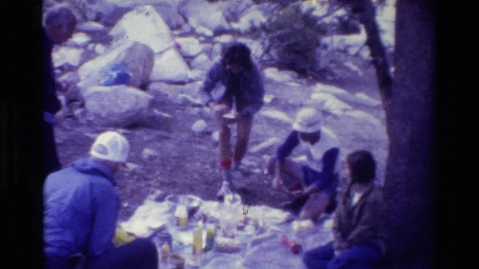 1981 home-movie frame of campers around a food blanket, with one person walking through the rocky campsite.